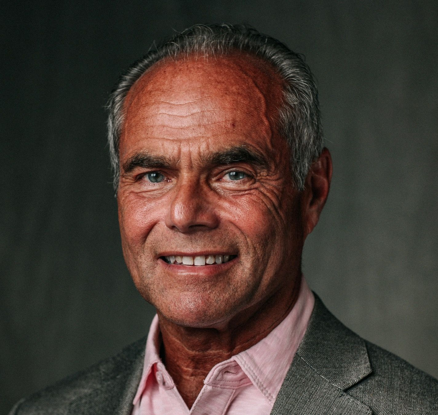 Man in gray blazer and pink shirt smiling; dark background.