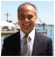 Smiling man in a suit outdoors by water; light skin, graying hair, tie, sunny background.
