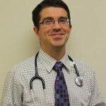 Doctor with stethoscope smiling, wearing glasses, checked shirt, and tie.