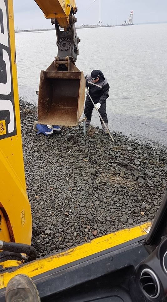 En mand står på en stenet strand ved siden af en gul gravemaskine.