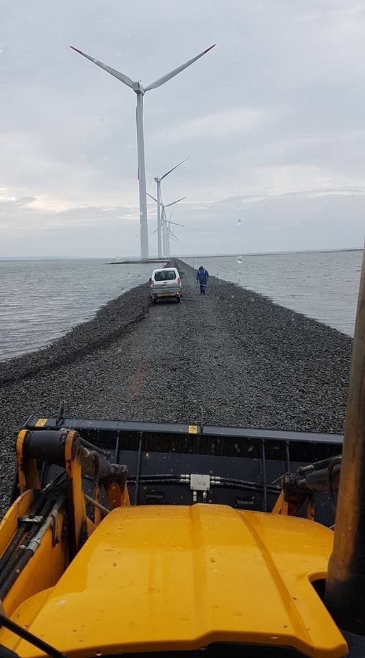 En bil kører ned ad en vej ved siden af ​​et vandområde med vindmøller i baggrunden.