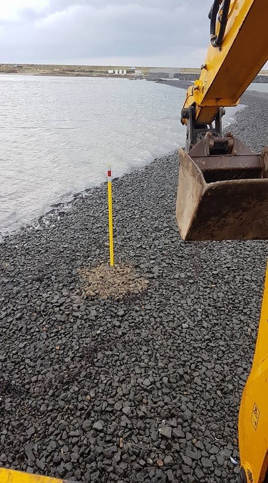 En gul gravemaskine graver et hul på en stenet strand ved siden af en vandmasse.