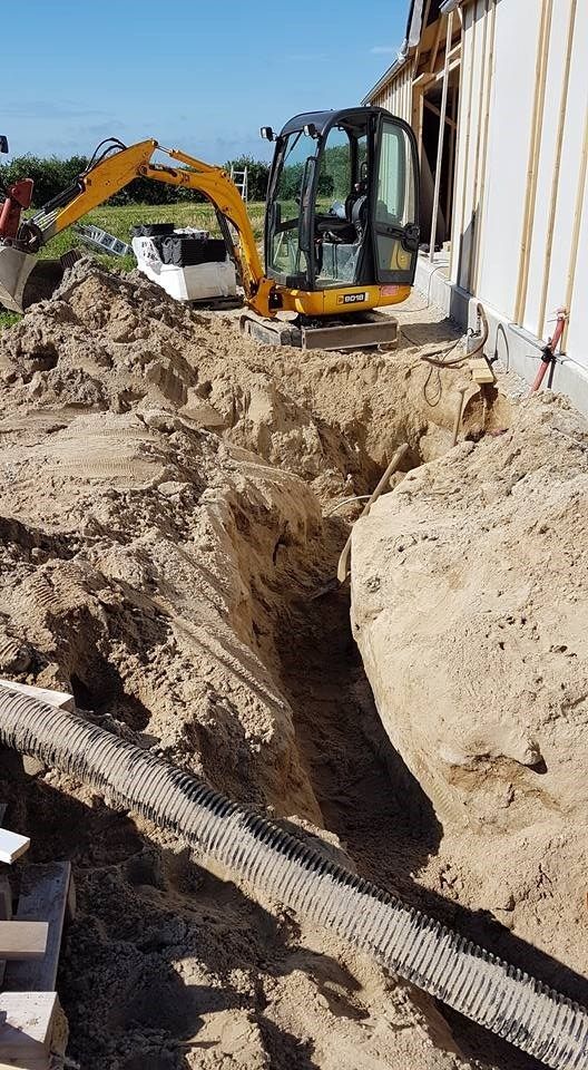 En gul gravemaskine graver et hul i snavset foran en bygning.