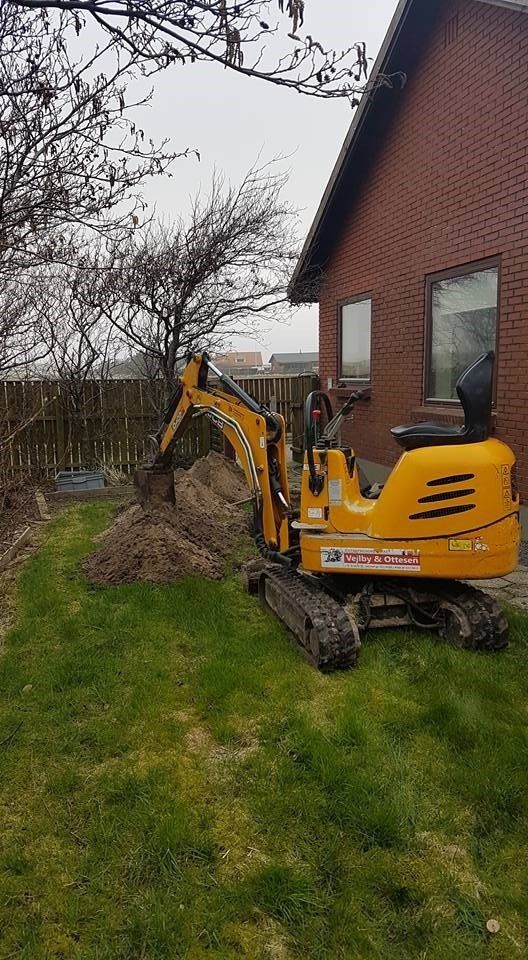 En lille gul gravemaskine sidder foran et murstenshus.