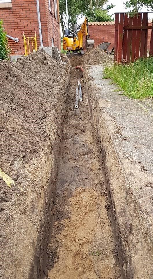 En gul gravemaskine graver en rende foran en murstensbygning.