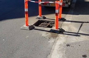 Et brønddæksel er omgivet af orange pæle i siden af vejen.