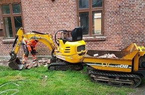 En mand arbejder på en lille gravemaskine foran en murstensbygning.