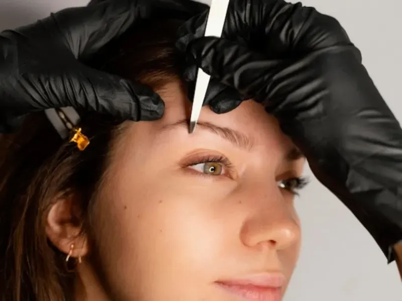 A woman is getting her eyebrows done by a person wearing black gloves.