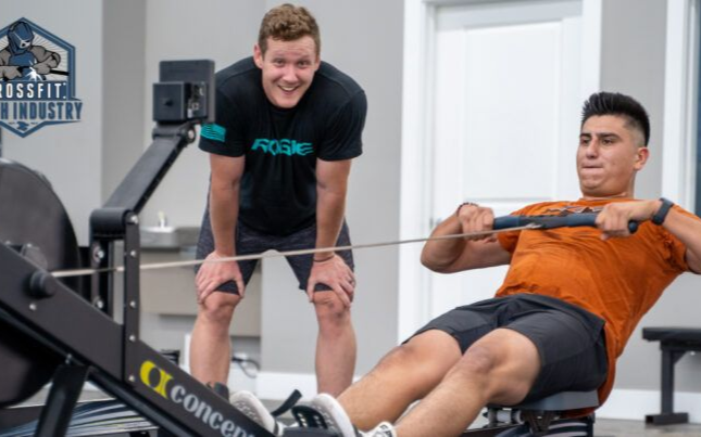 Man on rowing machine with coach watching in gym.