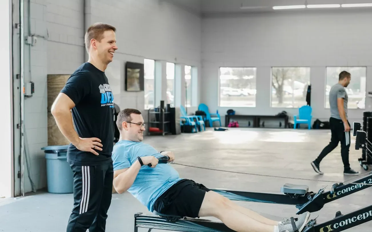 Man on rowing machine with trainer in a gym setting. Both smiling. Another person walks in the background.