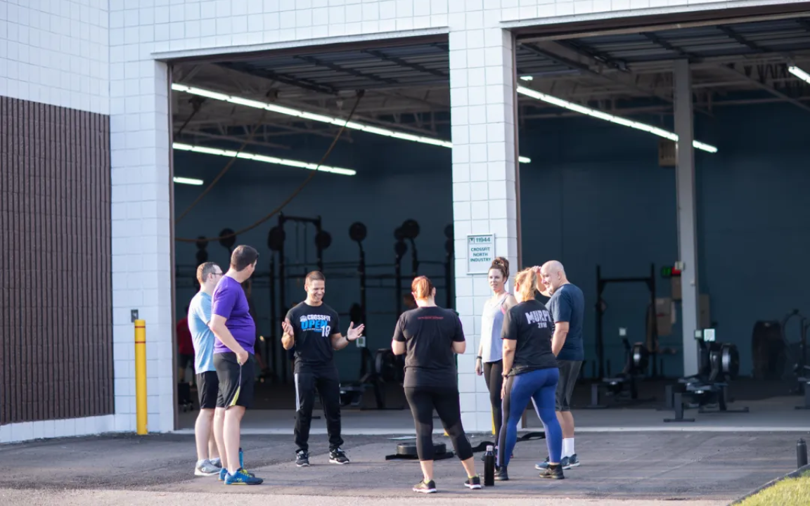 Group of people outside a gym, listening to a man. They are near open garage doors, in athletic wear.
