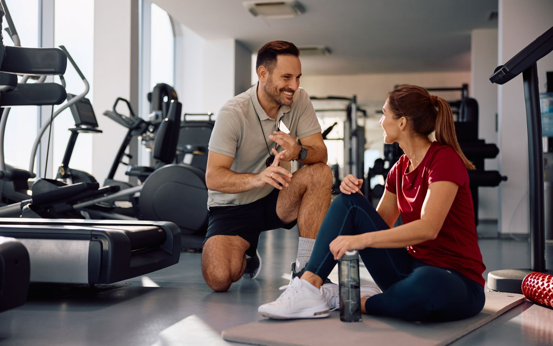 Man and woman talking in a gym, man kneeling, woman sitting, both smiling.