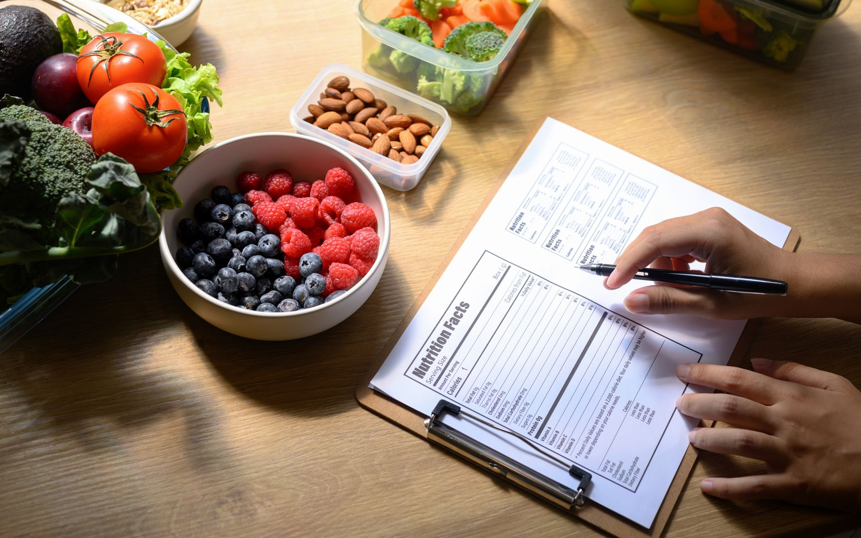 Person writing on a nutrition facts sheet, surrounded by fresh fruits, vegetables, and nuts on a wooden table.