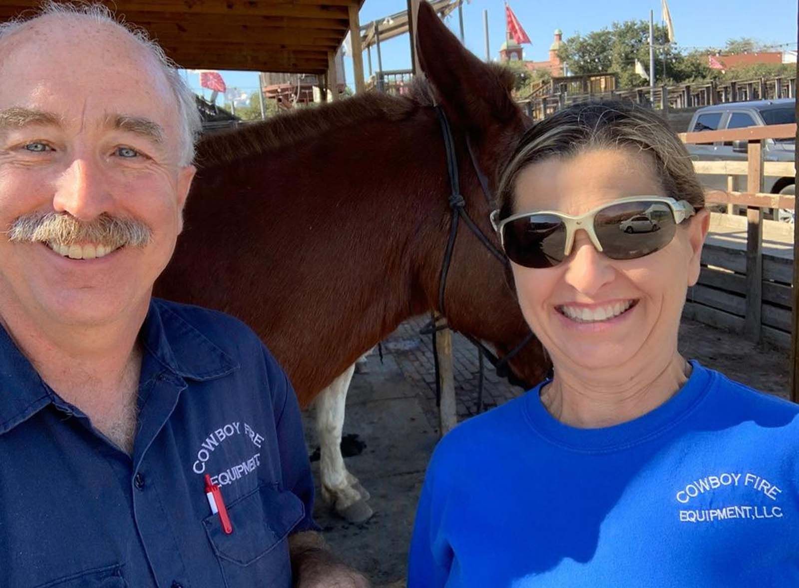 A man and a woman are posing for a picture with a horse in the background.