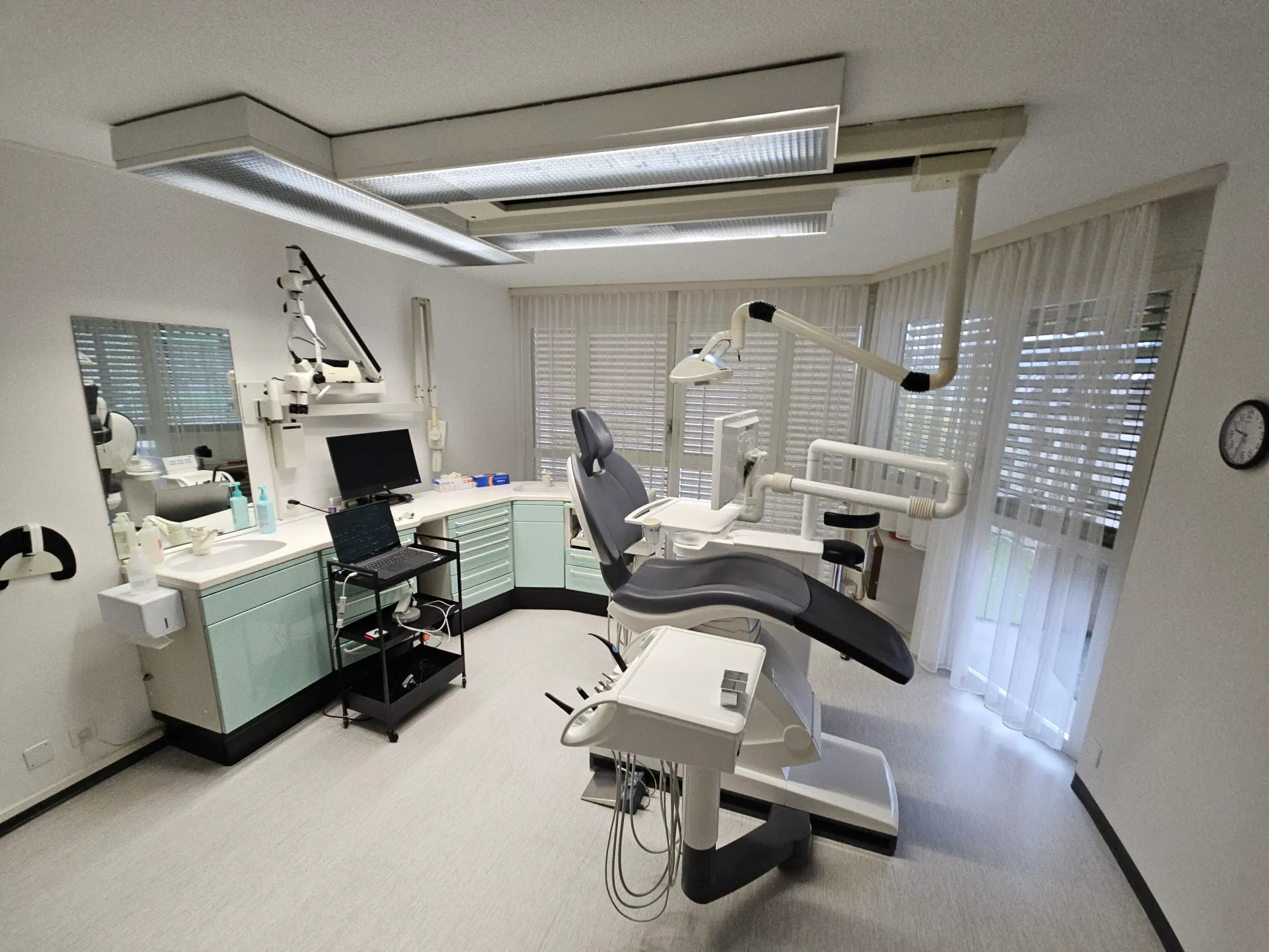 Une salle de soins dentaires avec un fauteuil pour le patient, du matériel médical, un comptoir et de grandes fenêtres avec stores.