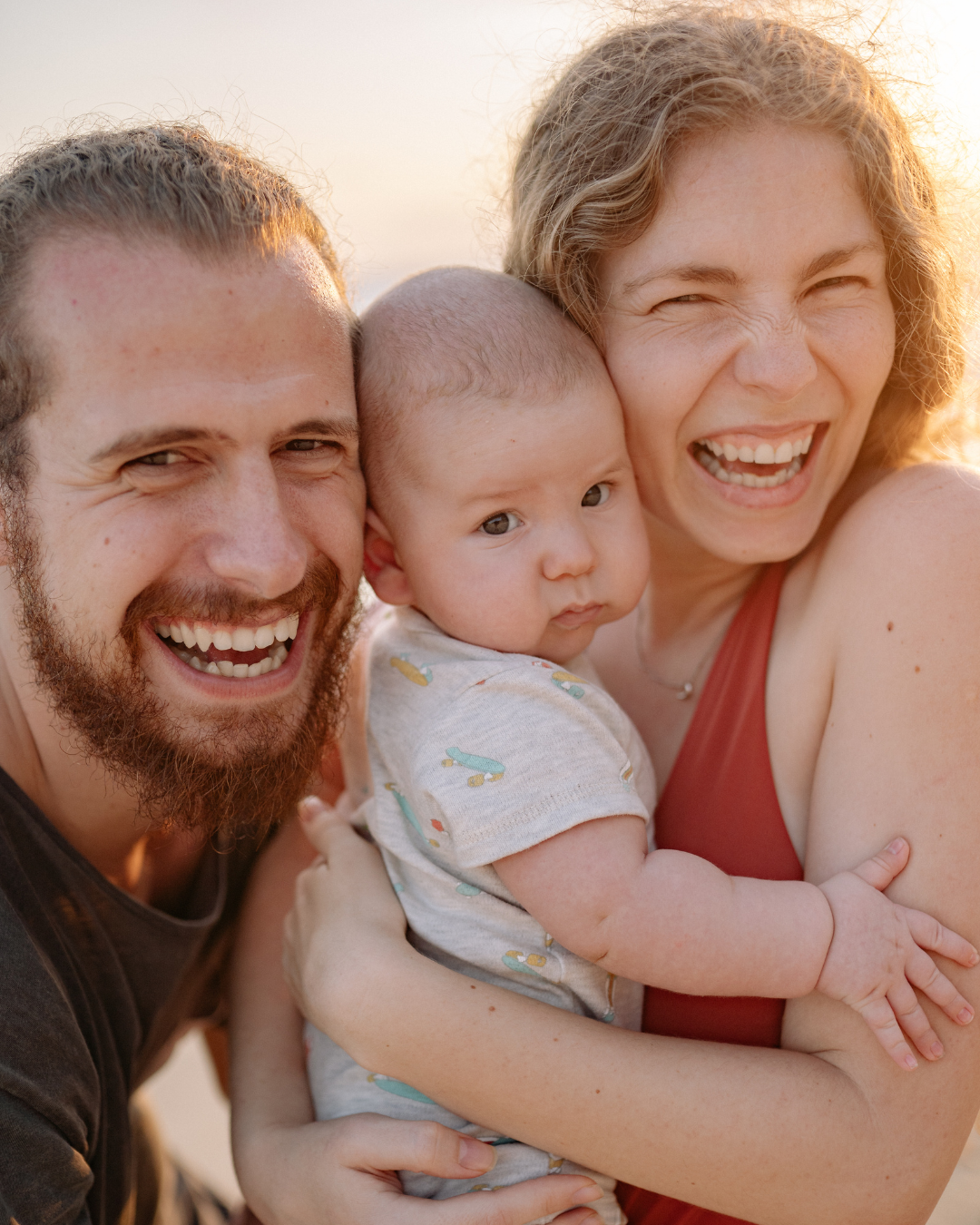 Un couple souriant pose pour une photo en gros plan à l'extérieur, tenant un bébé qui regarde l'objectif