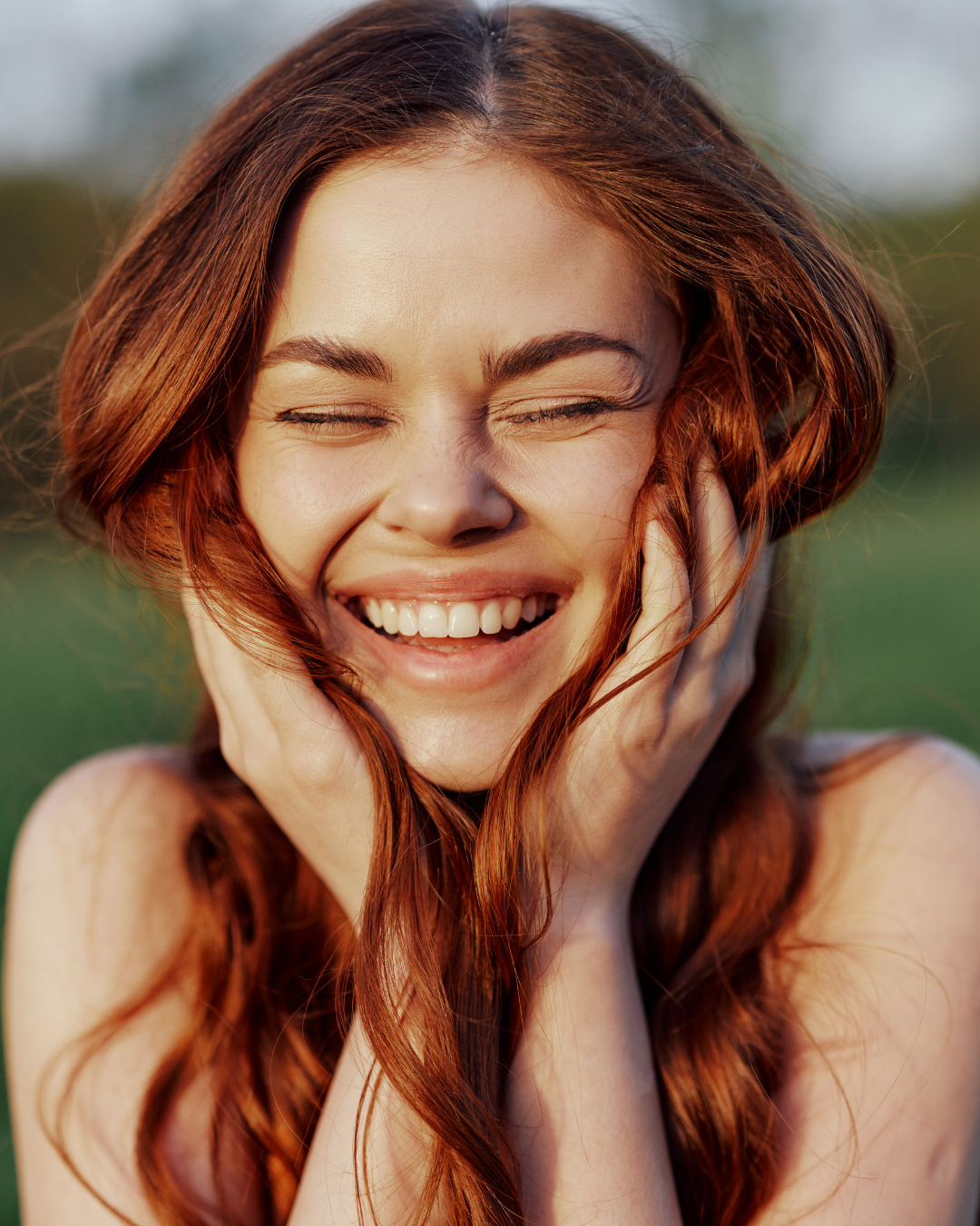 Une personne aux cheveux roux flamboyants sourit largement, les yeux fermés, les mains plaquées contre ses joues.