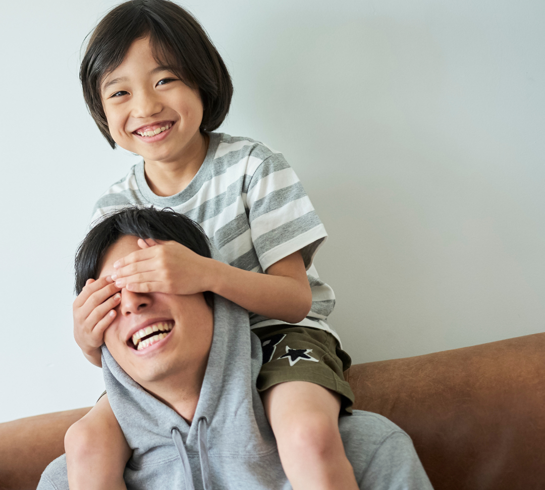 Une personne vêtue d'un sweat à capuche gris est assise sur un canapé tandis qu'un enfant souriant est assis sur ses épaules