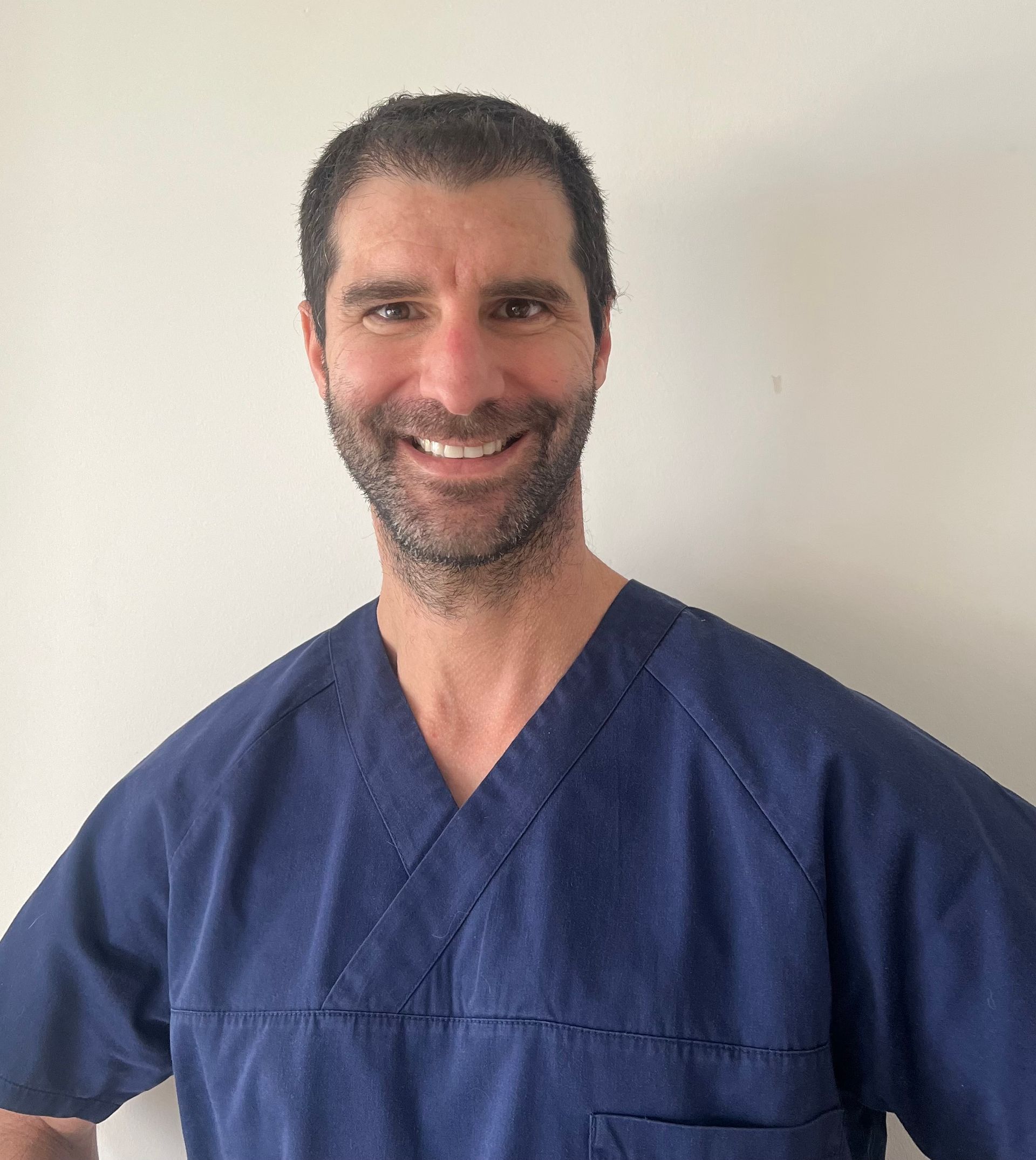 Une personne souriante, aux cheveux foncés et à la barbe fournie, portant une blouse médicale bleu marine à col en V