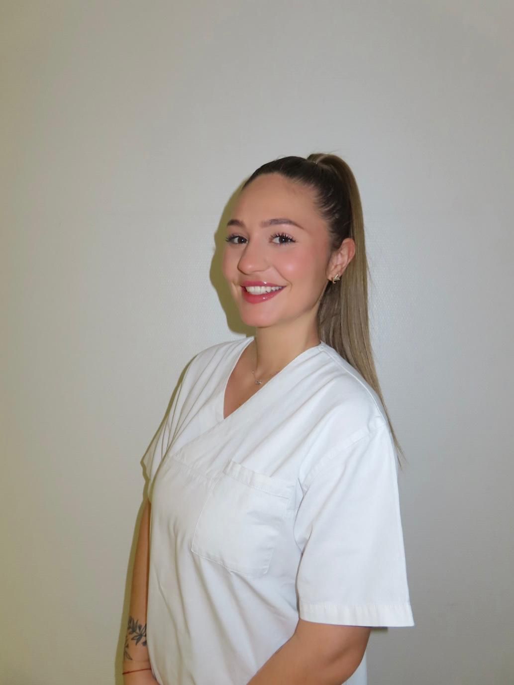 Une personne souriante aux longs cheveux attachés en queue de cheval, portant une tunique blanche à col en V