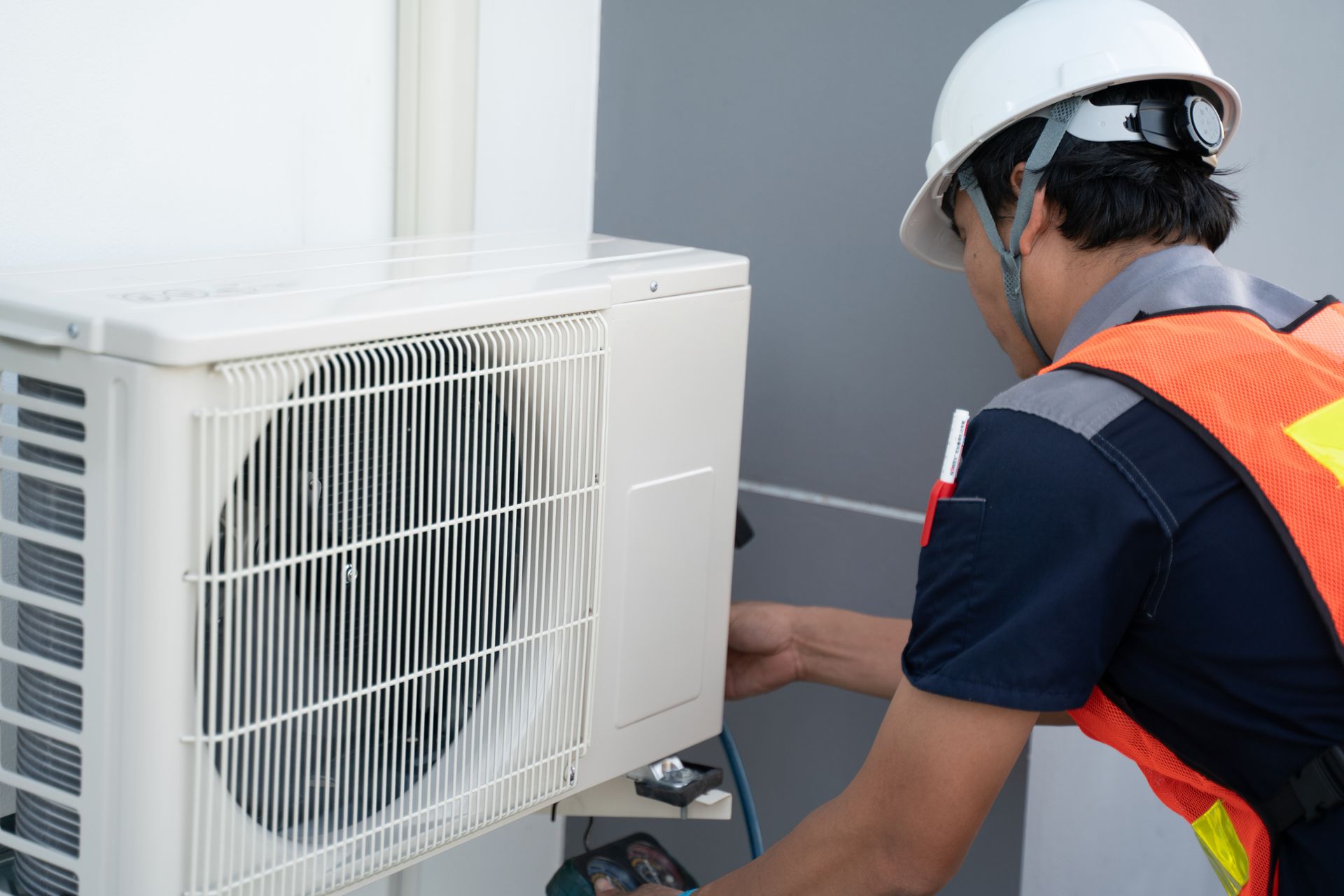 An HVAC technician in a safety vest is working on a white air conditioner