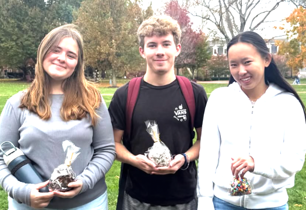 Three people stand outdoors on a grassy lawn, each holding a small, clear bag of candy.