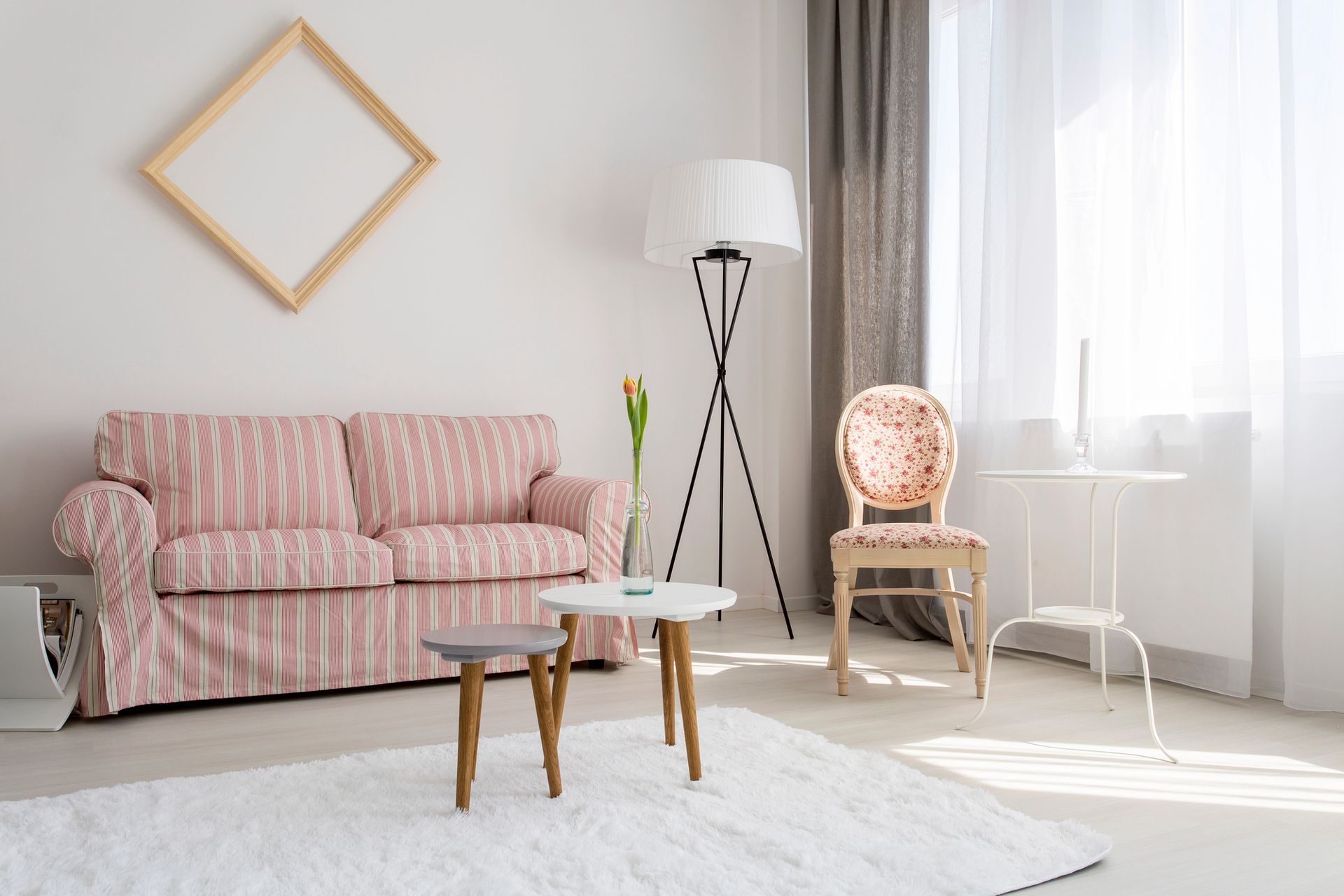 Sala de estar luminosa con un sofá rosa, mesitas, lámpara de pie, silla y luz natural que entra por la ventana.