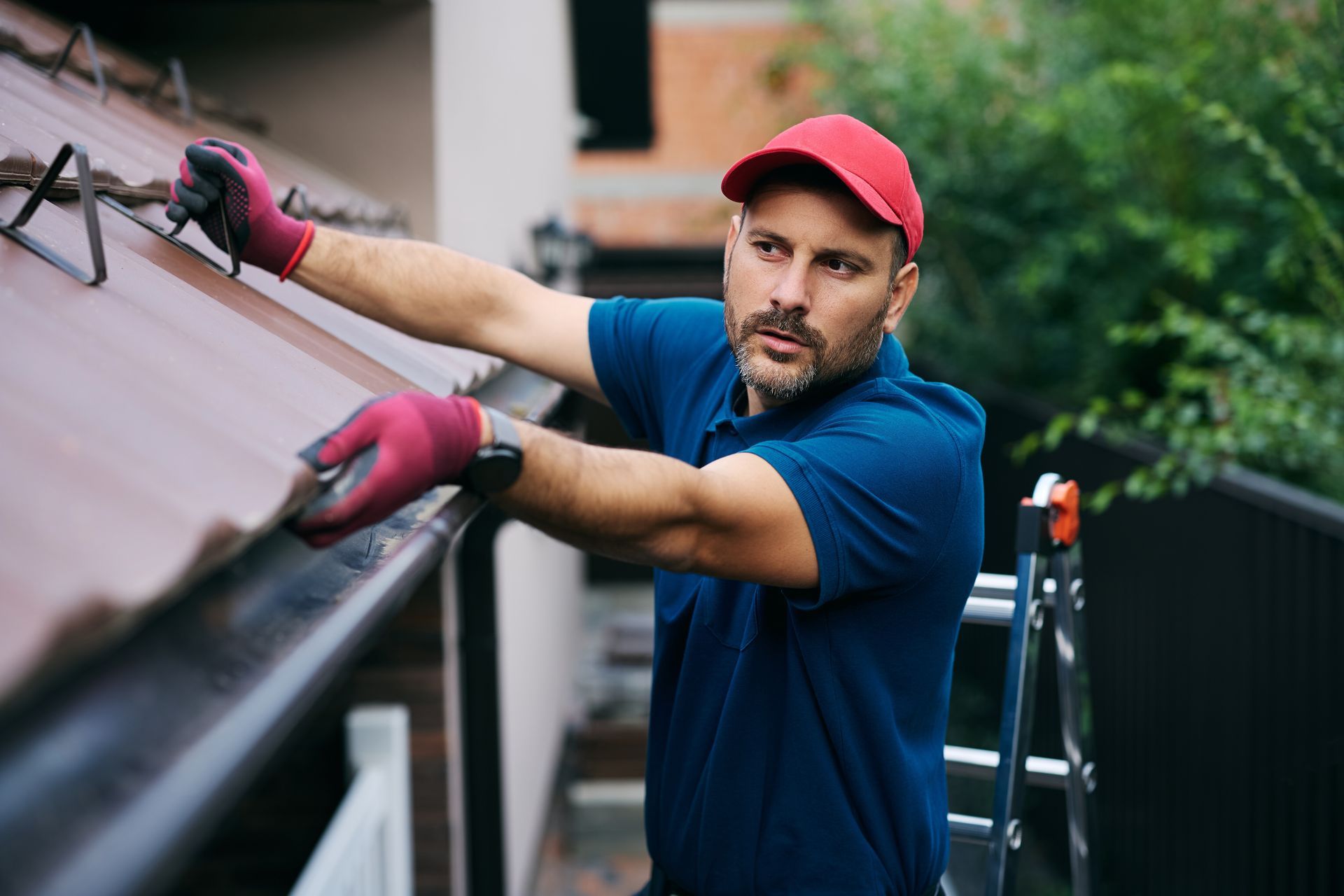 A worker in a blue shirt and red cap wearing protective gloves inspects roof gutters from a ladder.