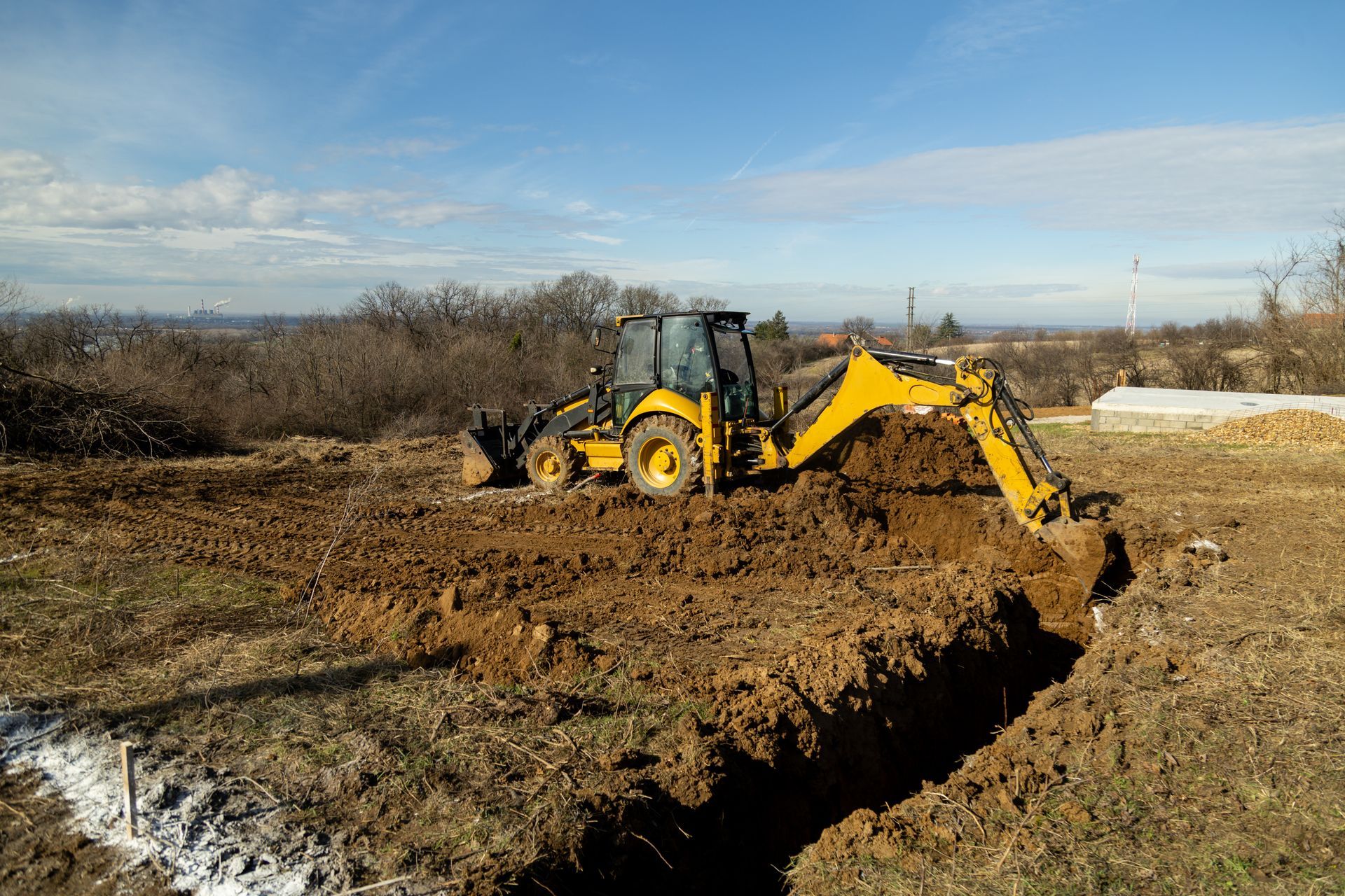 A yellow backhoe excavator digs a trench in a field on a sunny day.