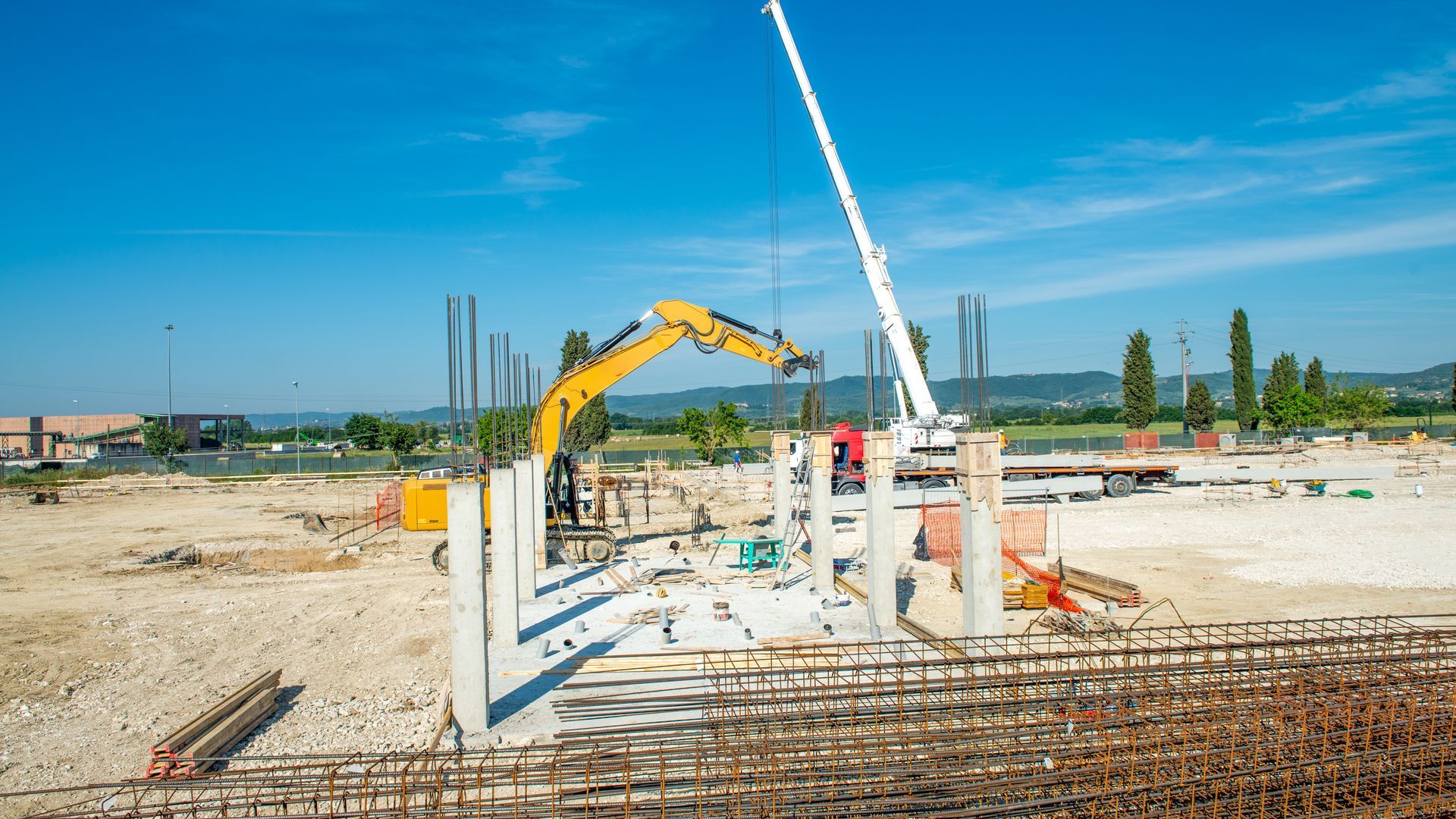 Construction site with an excavator and a crane operating near vertical concrete pillars under a clear blue sky.