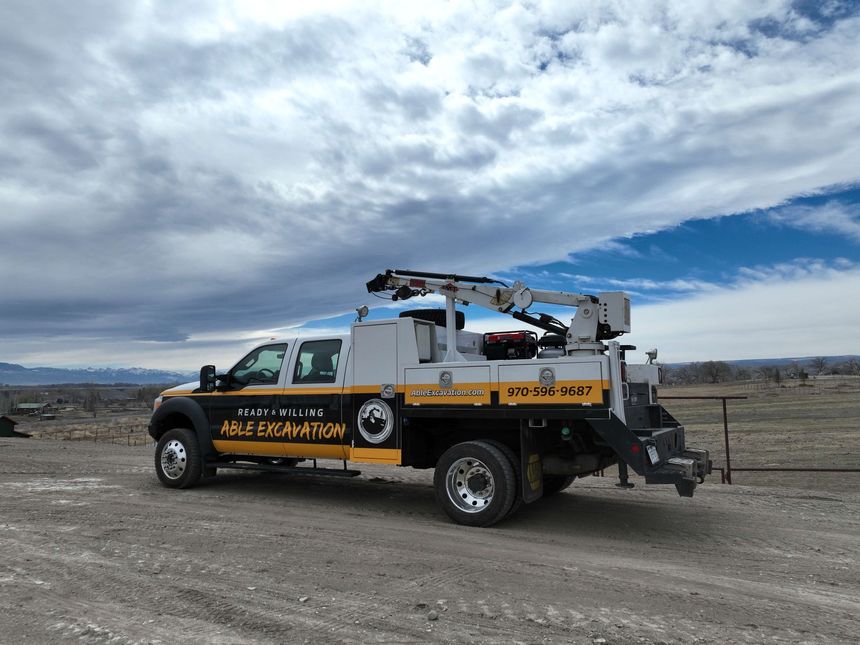 An Able Excavation yellow and black utility truck with an extendable boom crane parked on a dirt lot under a cloudy, blue sky near Delta, CO.