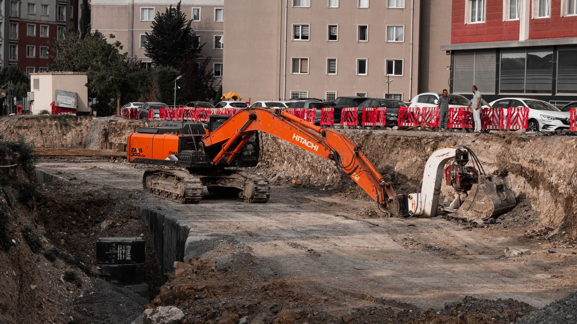 Orange excavator in a deep construction pit with apartment buildings and red barriers in the background