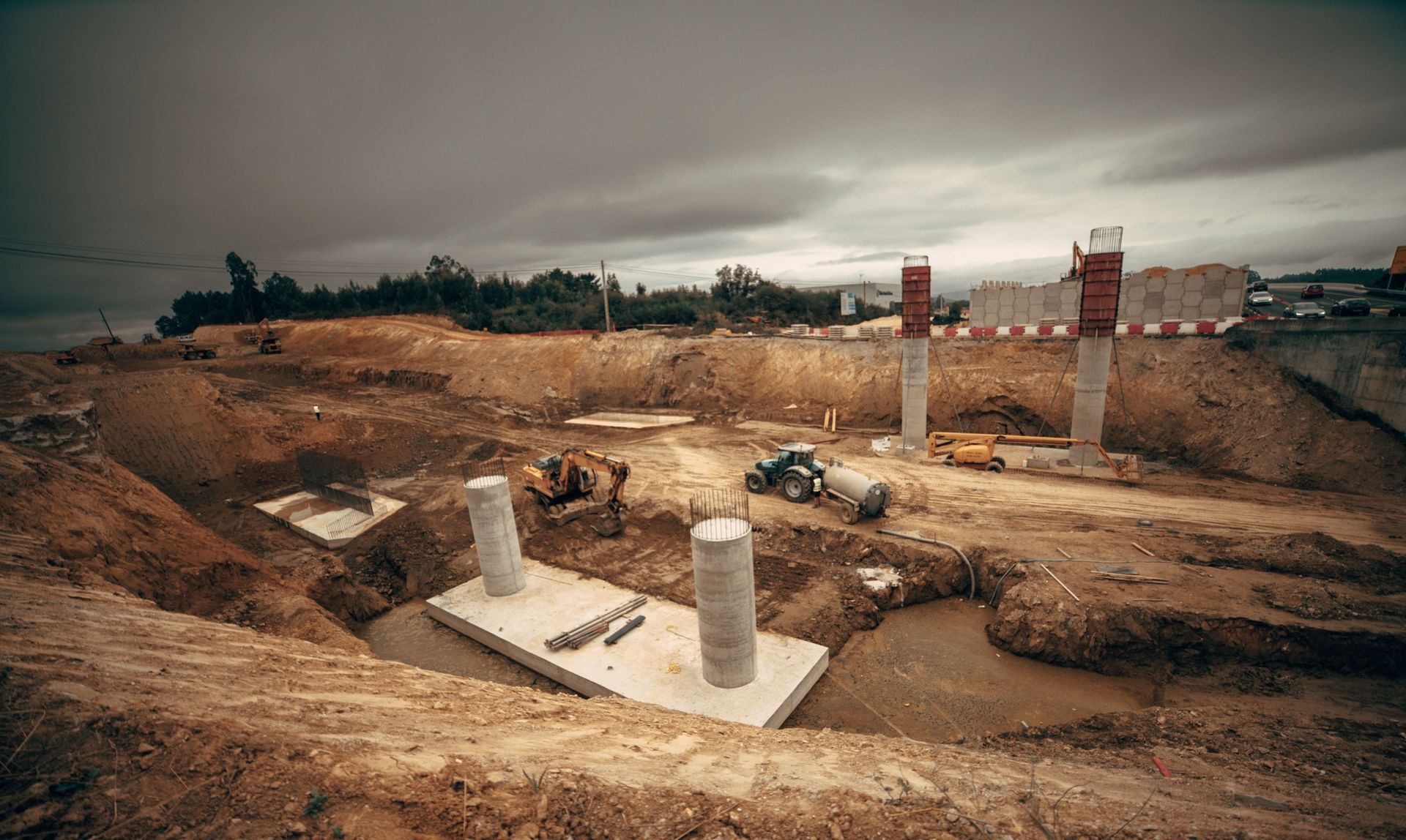 Archaeological excavation site with concrete pillars, dirt pits, and overcast sky
