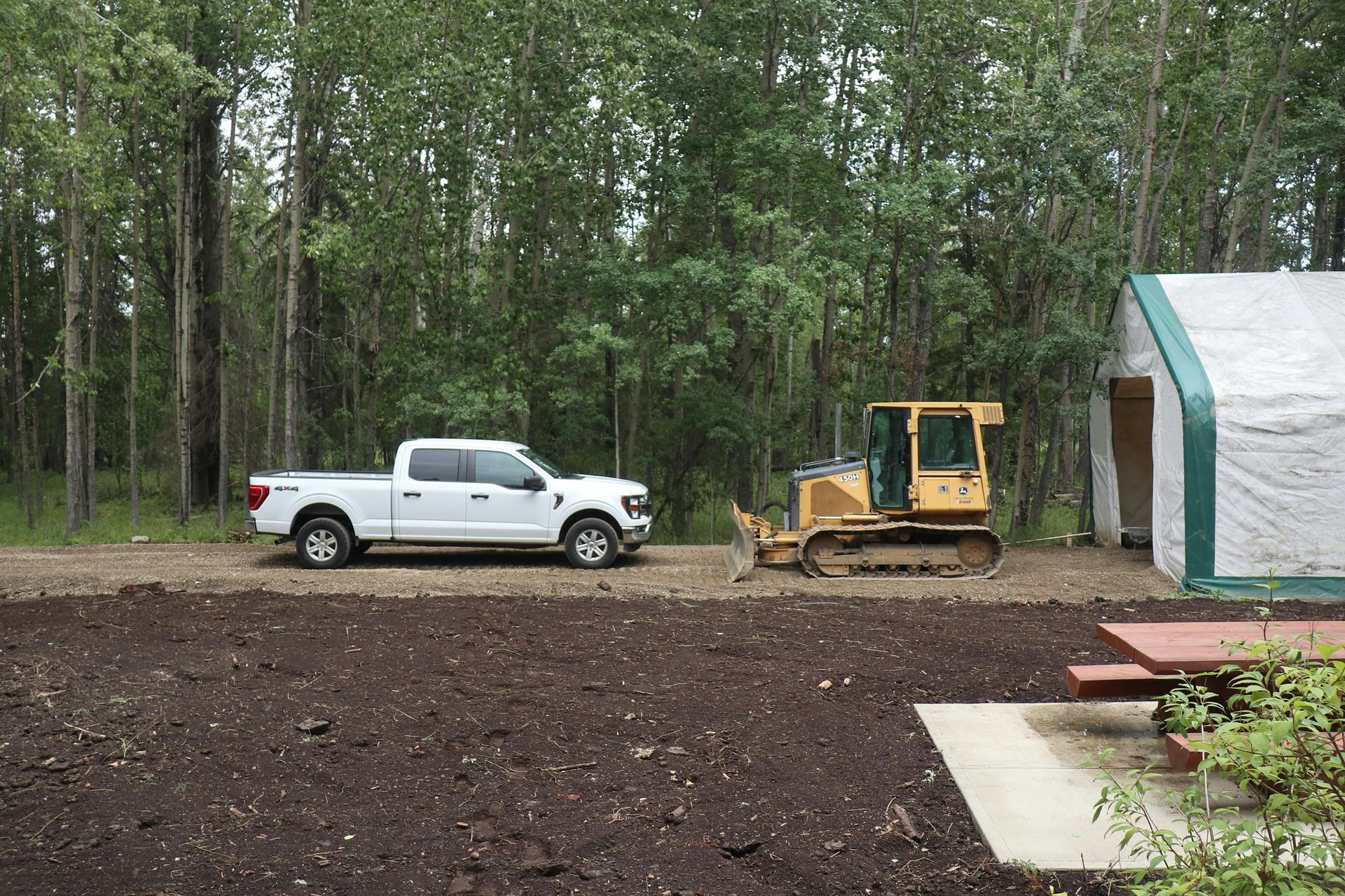 White pickup and yellow bulldozer parked beside a shed in a wooded yard after rain