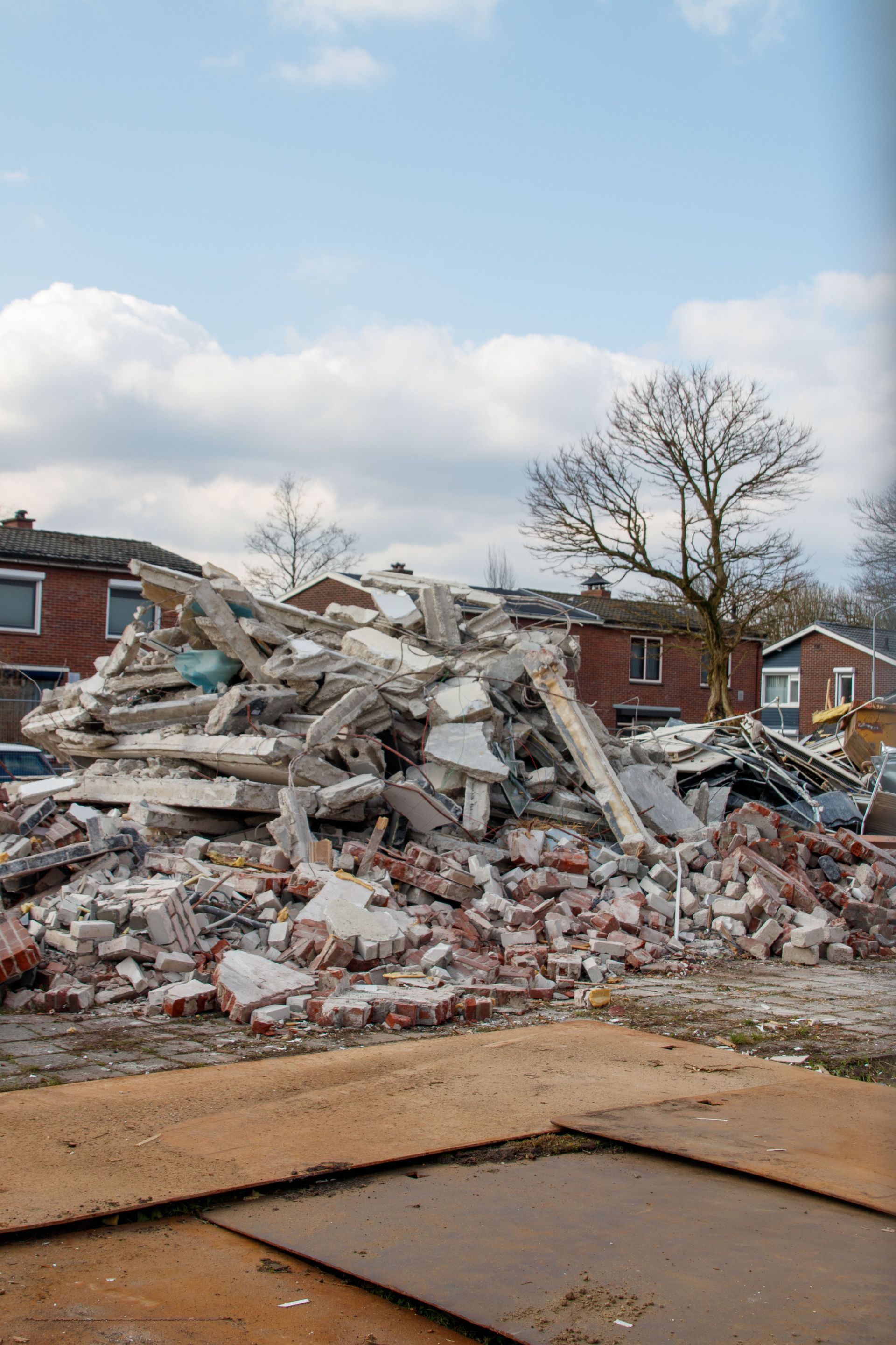 Collapsed brick building rubble in a residential neighborhood under a cloudy sky