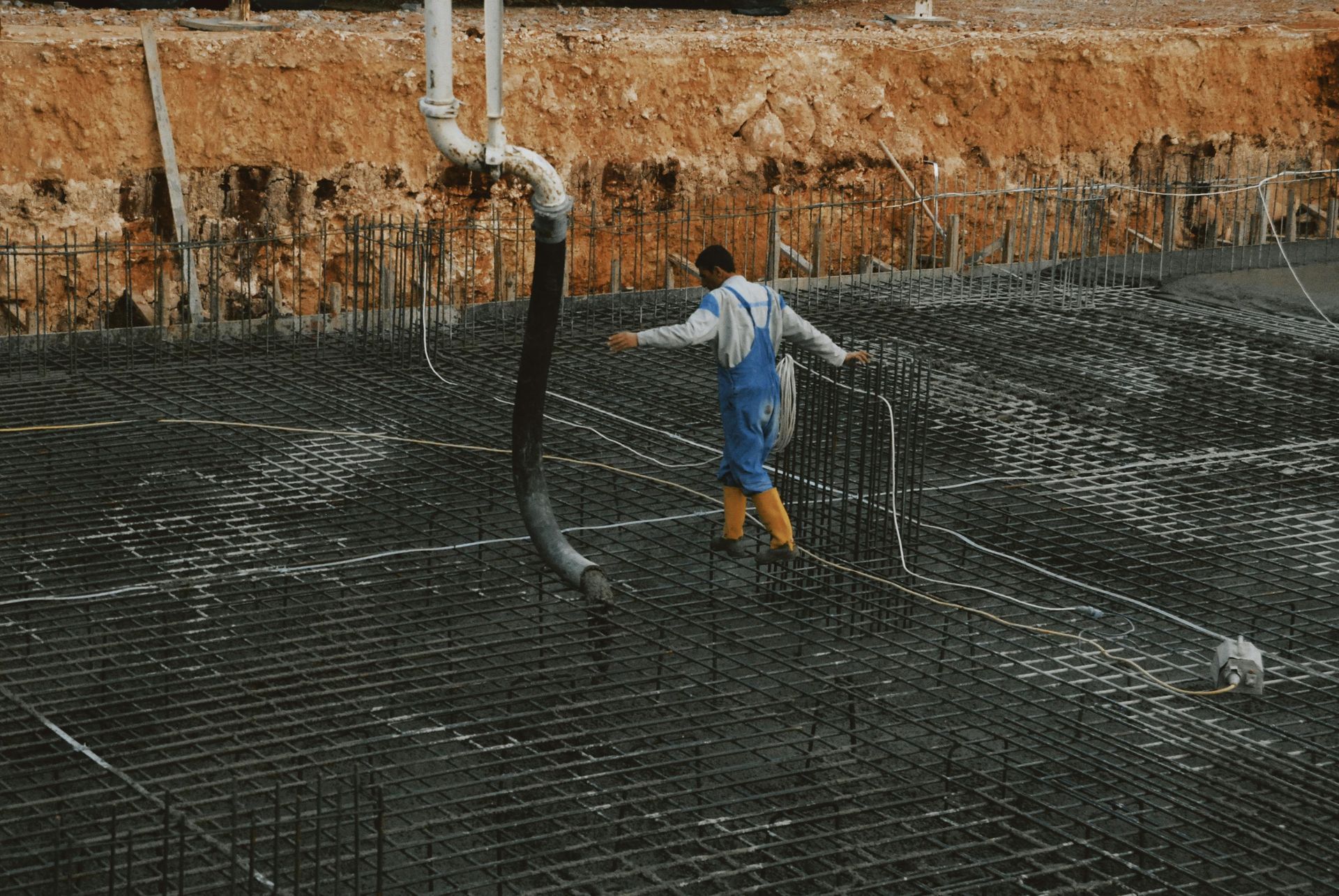 Worker in blue overalls and yellow boots in a wet concrete pit with rebar mesh and pipes.