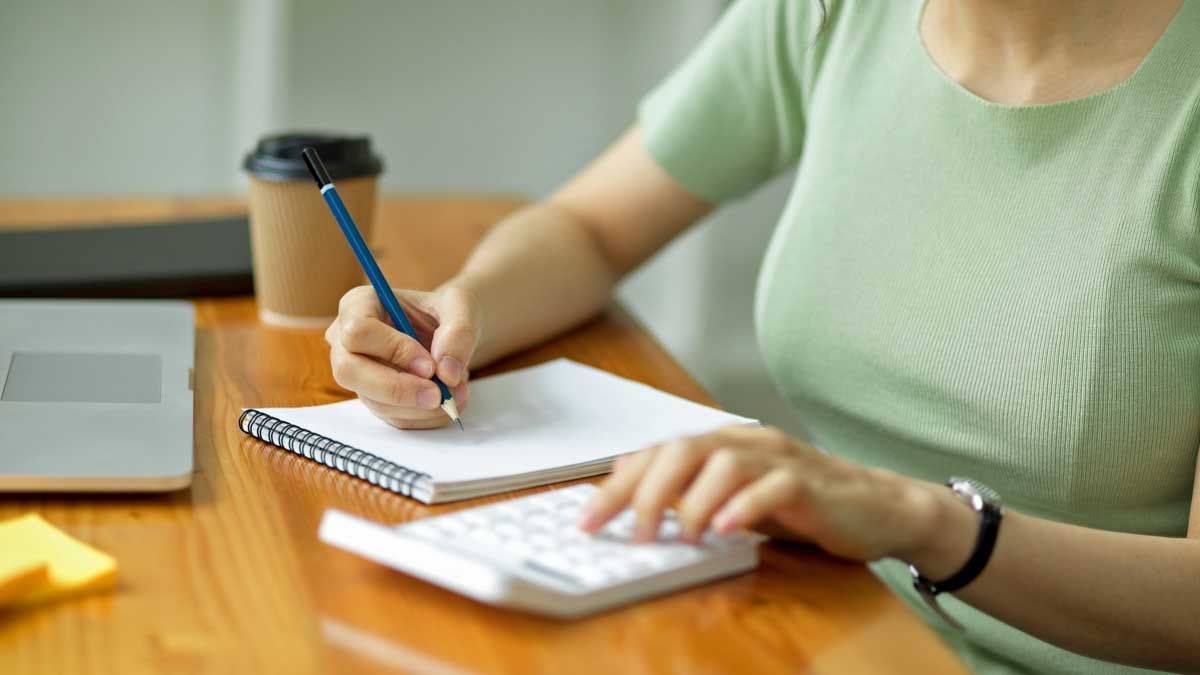 Person writing in a notebook, using a calculator, and holding a pencil. Beside them is a coffee cup and a laptop.