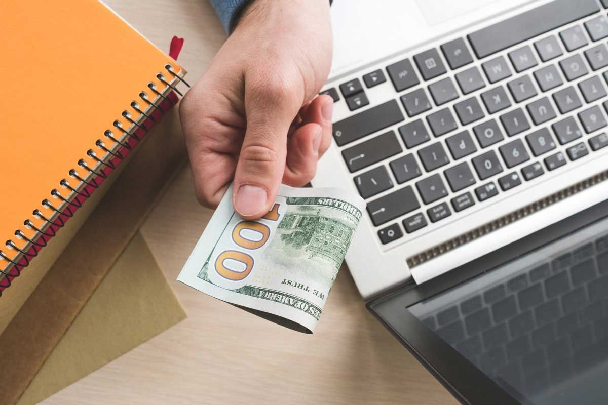 Hand holding a $100 bill over a laptop next to a stack of notebooks.