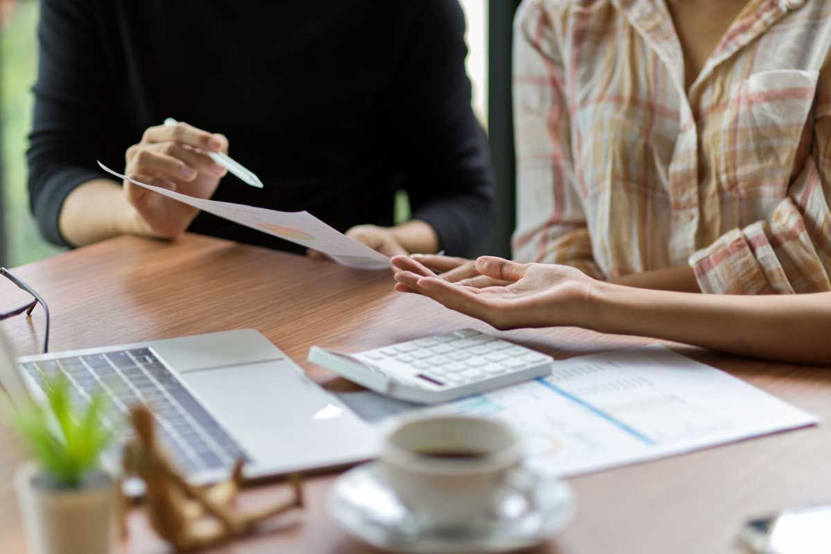 Two people reviewing documents at a table with a laptop, calculator, and coffee.
