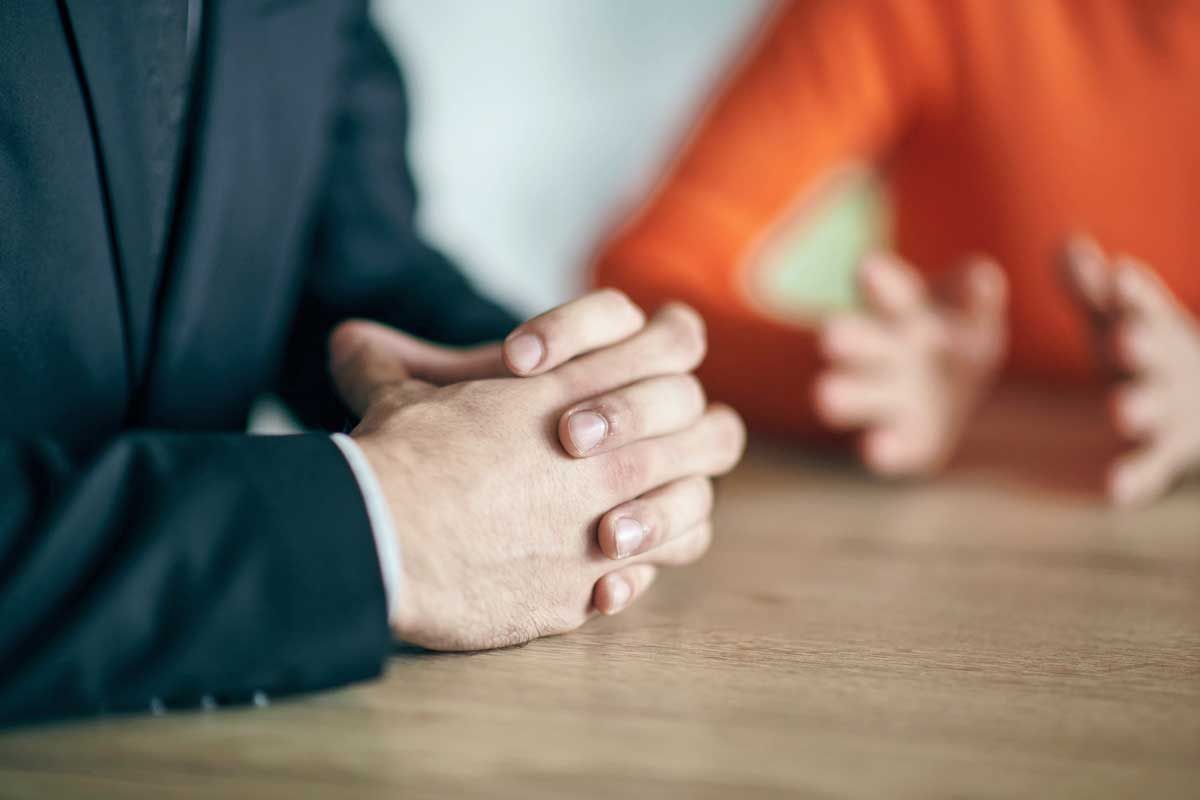 Person in suit with hands clasped on table, conversing with someone in orange sweater.