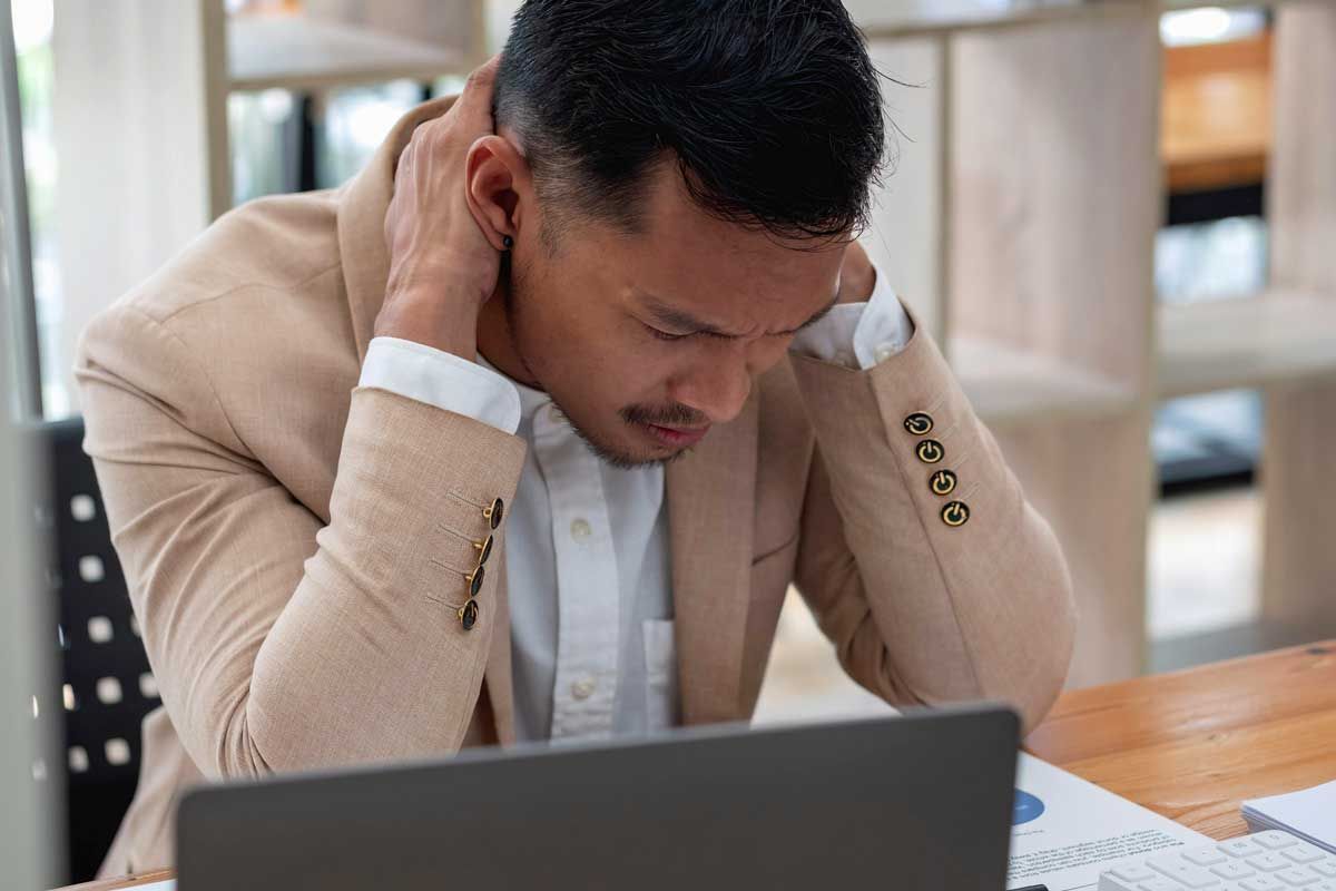 Man in tan blazer, holding neck, looking down, possibly stressed, at laptop in office setting.
