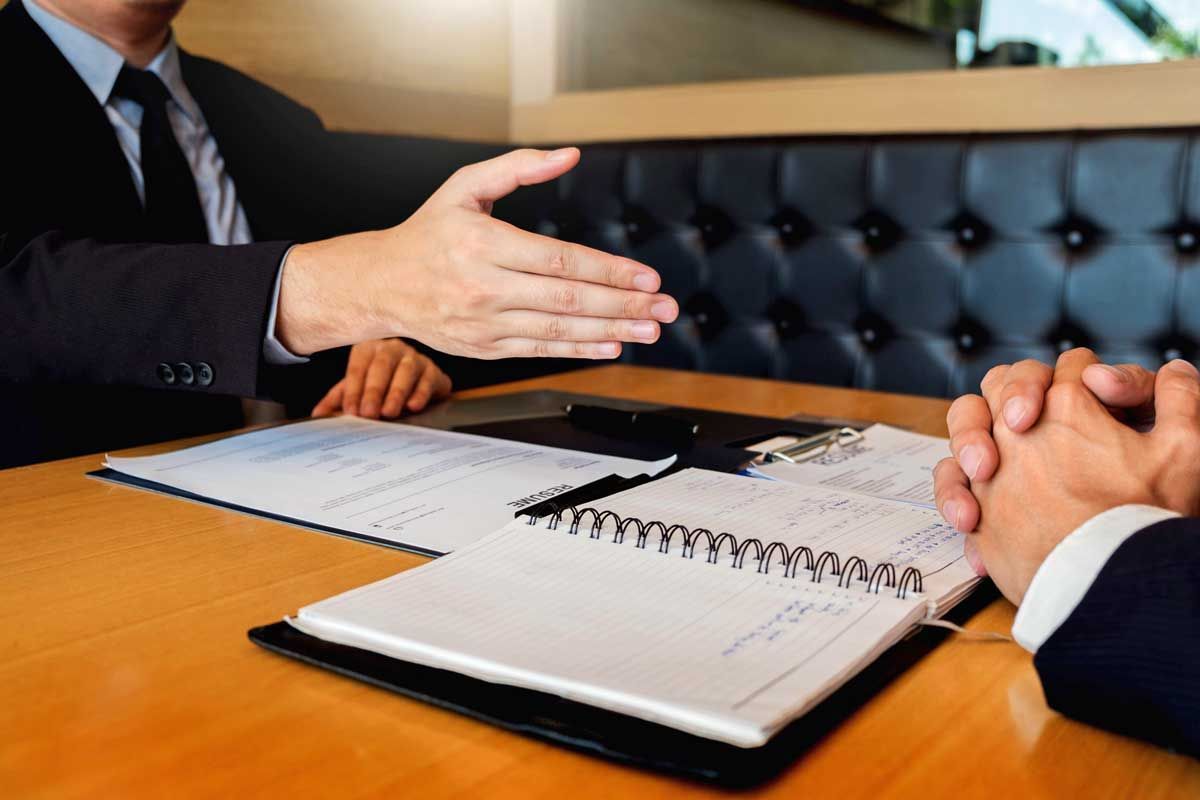 Two men in suits at a table, one extending a handshake, paperwork visible.