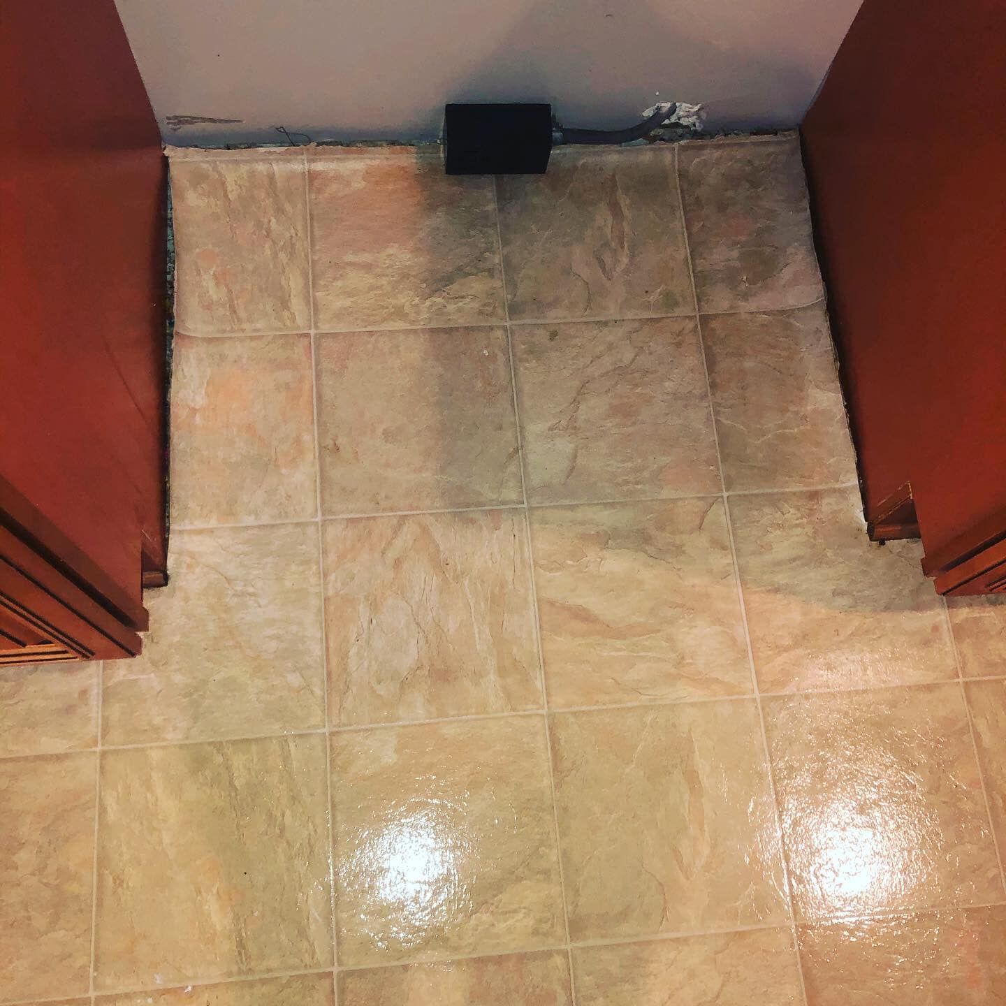 A kitchen floor with tan, mottled square tiles between two wooden cabinets and a small black electrical box on the wall.