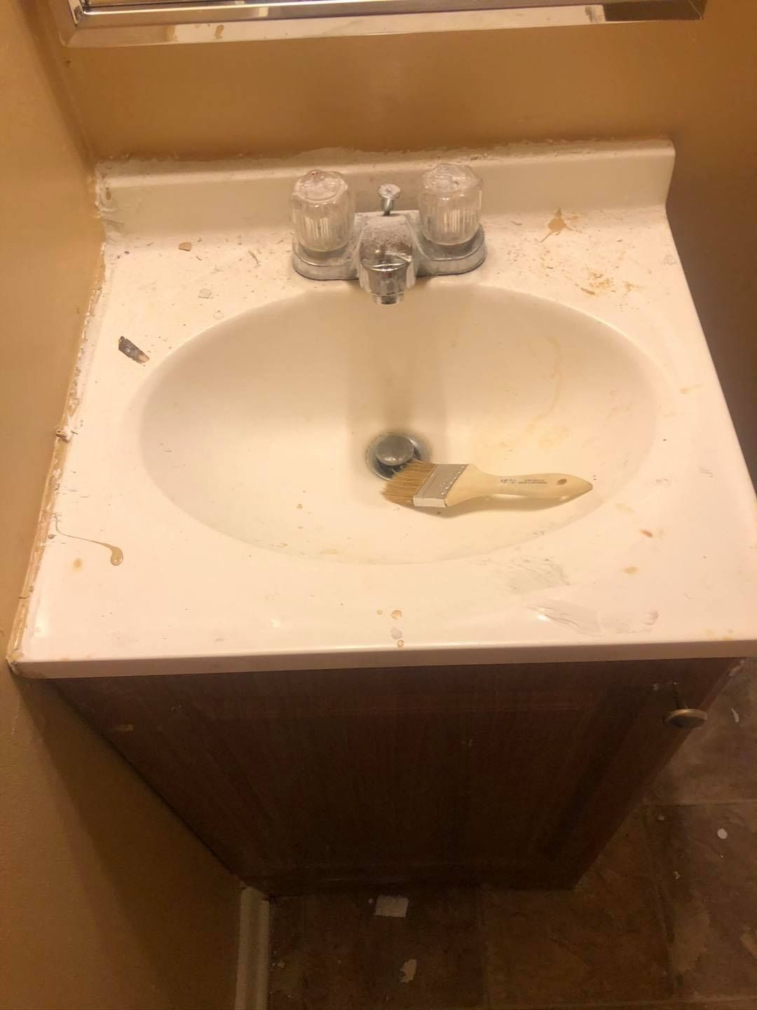 A white bathroom sink cabinet with a paint-splattered countertop and a paintbrush resting in the basin.
