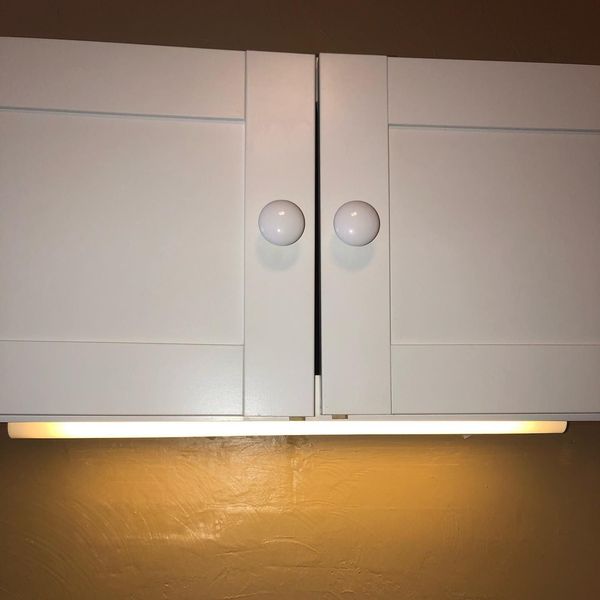 A pair of white kitchen cabinets with round knobs and an under-cabinet light strip glowing against a dark yellow wall.