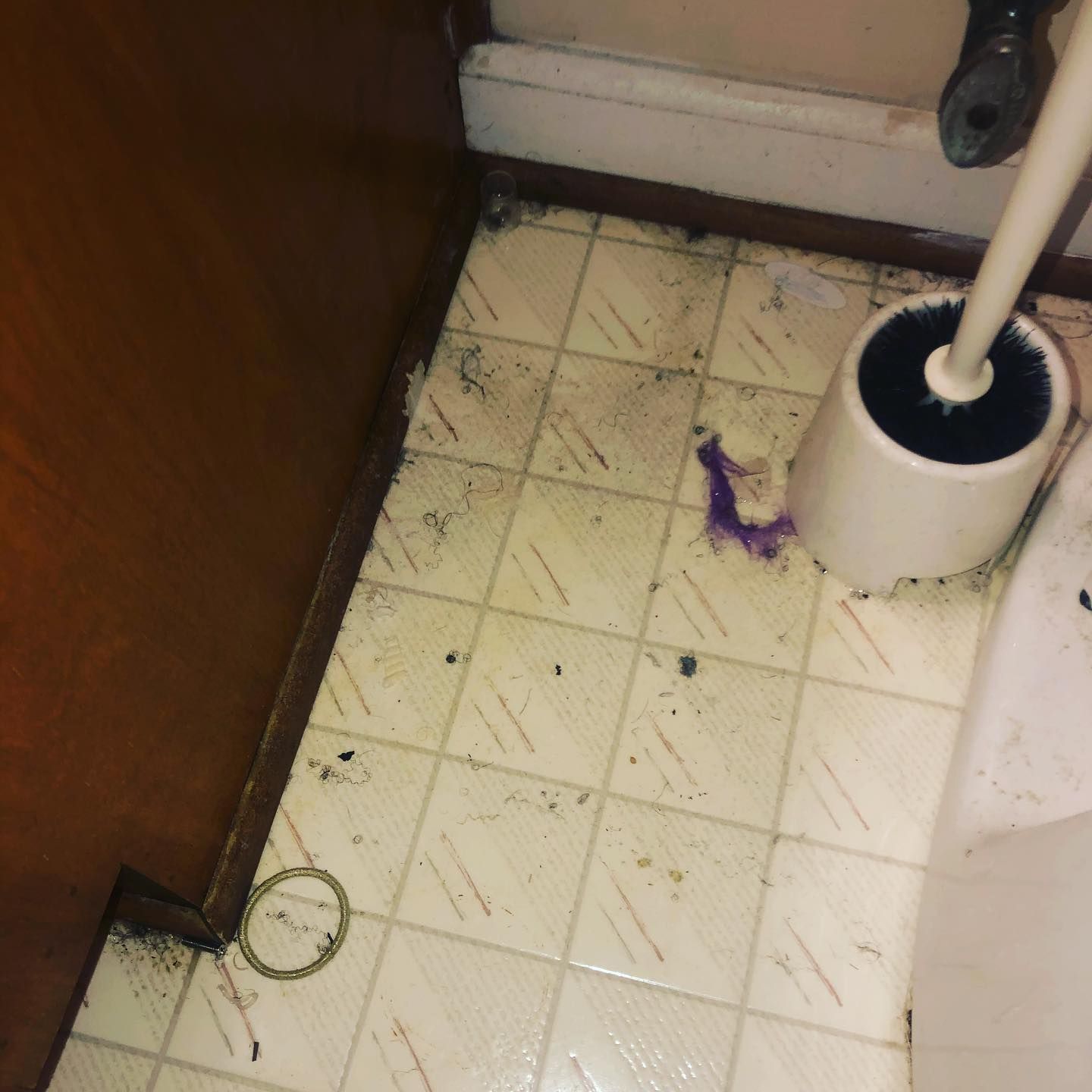Dirty bathroom floor tiles with debris, a toilet brush holder, and a stain near a wooden cabinet base.
