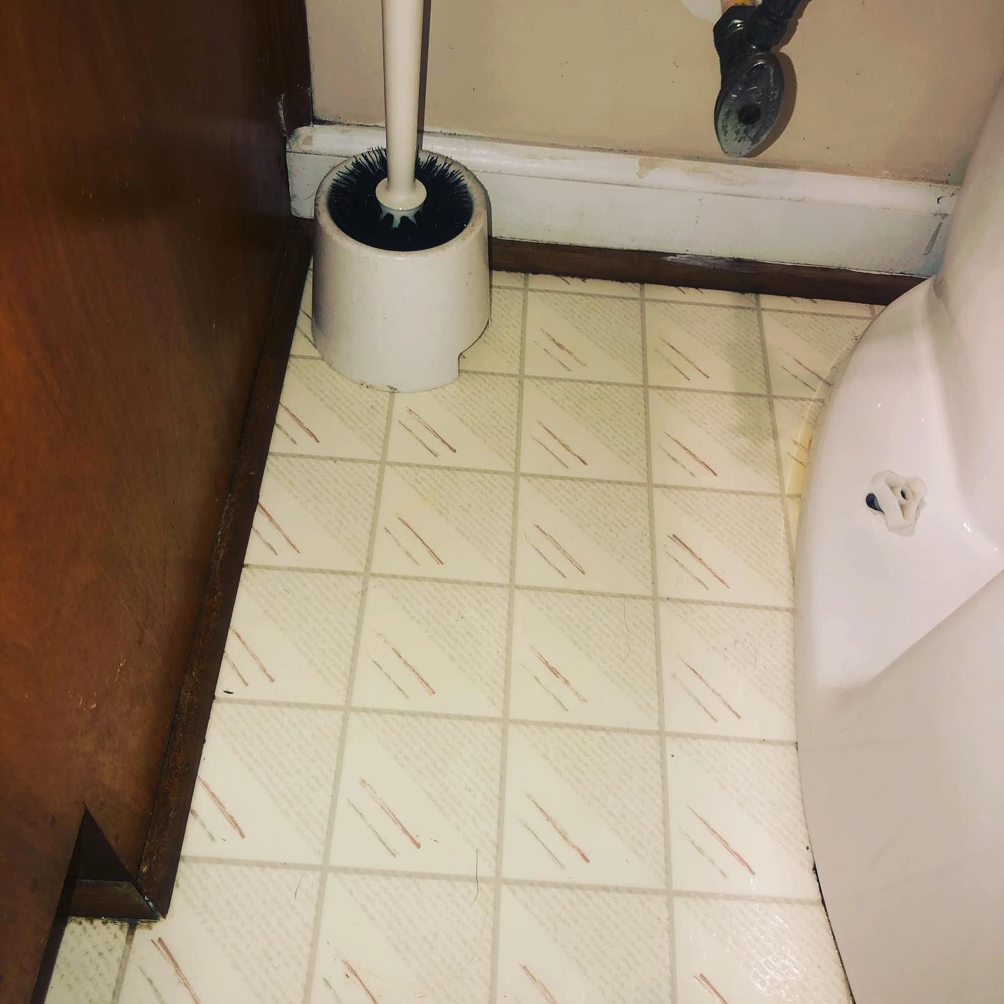 A white toilet brush holder sits on a light-colored, square-tiled bathroom floor between a dark cabinet and a white toilet.