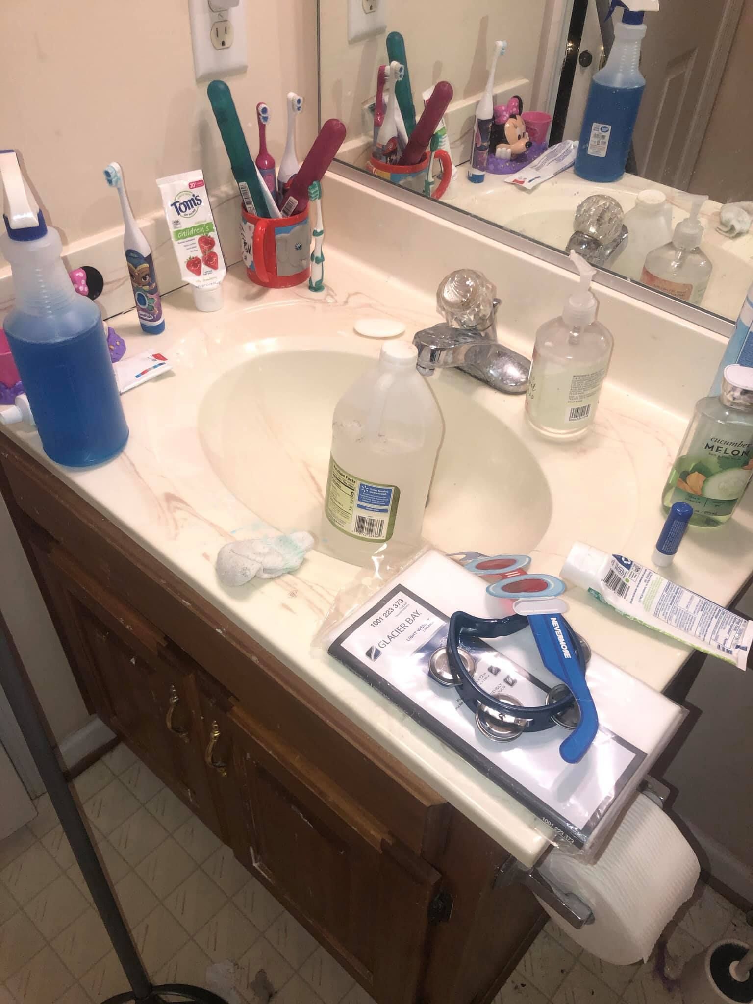 A cluttered bathroom sink counter with toothbrushes, cleaning sprays, a white plastic jug, and various personal items.