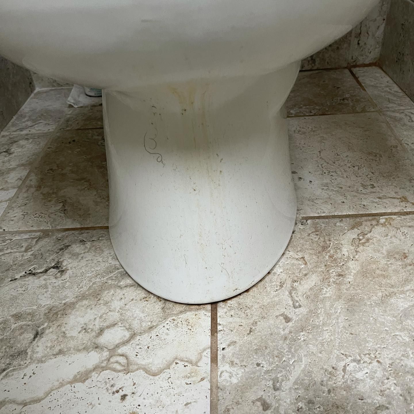 Close-up of a white toilet base with brown liquid stains running down the porcelain onto tan stone floor tiles.