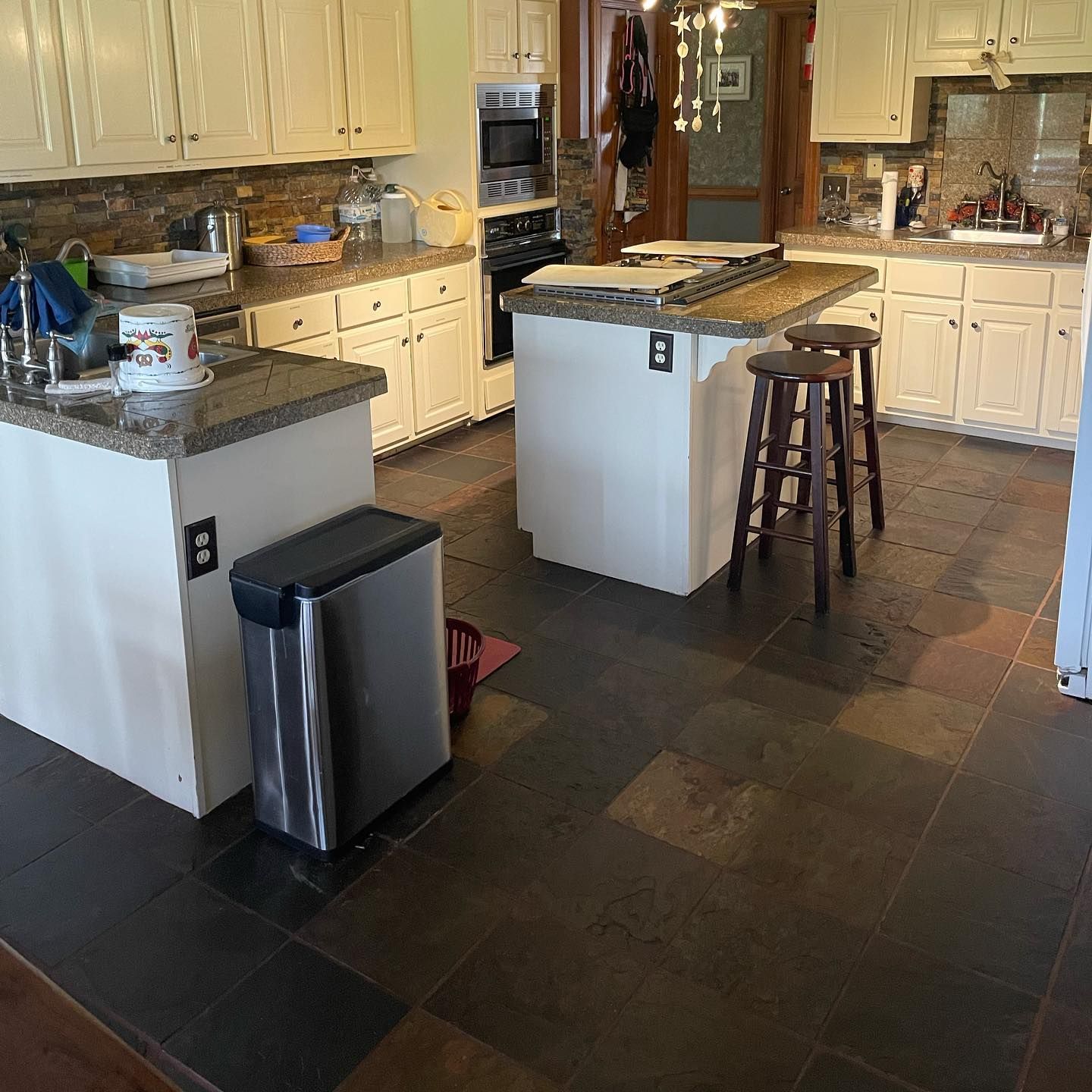 A spacious kitchen with white cabinets, stone countertops, a central island with two stools, and dark tiled flooring.